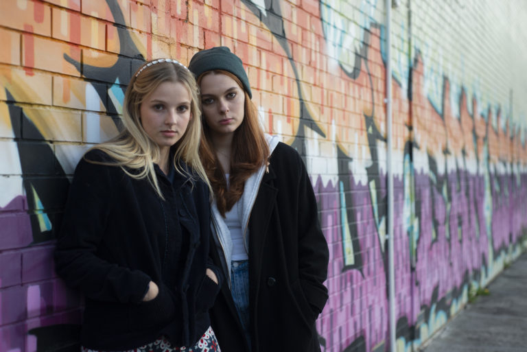 Heidi (Madelyn Sheahan) and Tabby (Georgia Crisfield-Smith) lean against a graffiti wall (landscape)